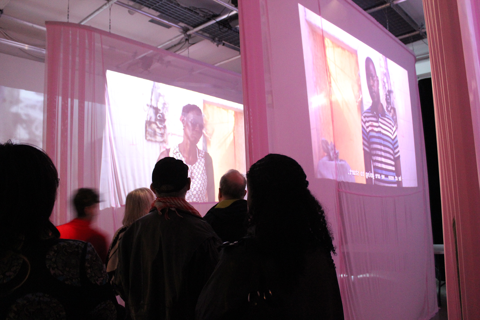 Rendition Exhibition Opening Video projected onto pink curtains, with shadows of people gathered around at the opening exhibition of Rendition